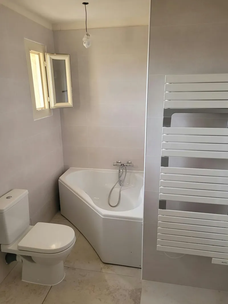Salle de bain avec baignoire d’angle, fenêtre et grand sèche-serviettes blanc